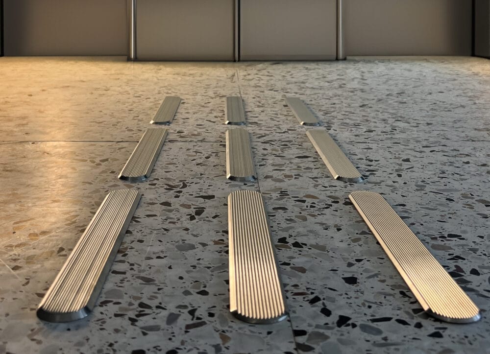 Taktila ledstråk i rostfritt stål med räfflad struktur installerade i ett terrazzogolv. Ledstavarna bildar tre parallella linjer.