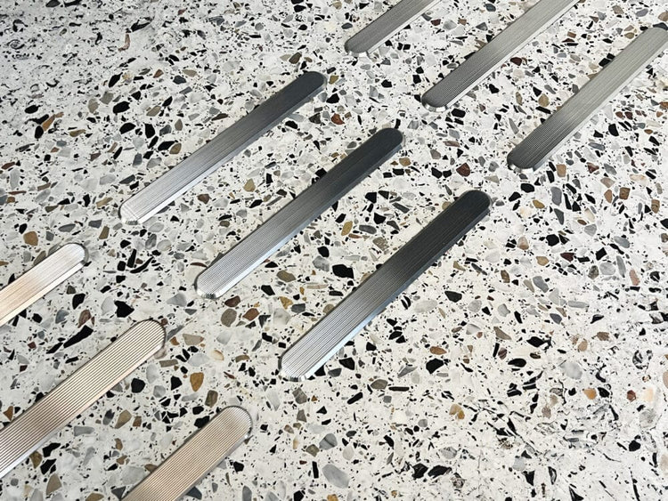 Taktila ledstråk i rostfritt stål monterade i ett terrazzogolv med vit bas och mörka inslag. Ledstavarna har en räfflad yta och skapar ett mönster som leder besökare genom utrymmet.