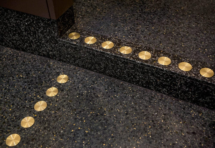 Taktila markeringar i mässing bildar ledstråk längs trappkant och golv på mörk terrazzobotten.
