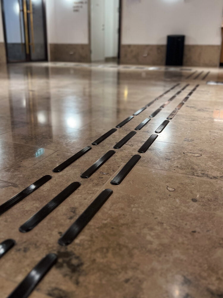 Taktila ledstråk från Safestep SVARTBÄCKEN med svarta PVD-belagda stavar som kontrasterar mot ljusbrunt stengolv.