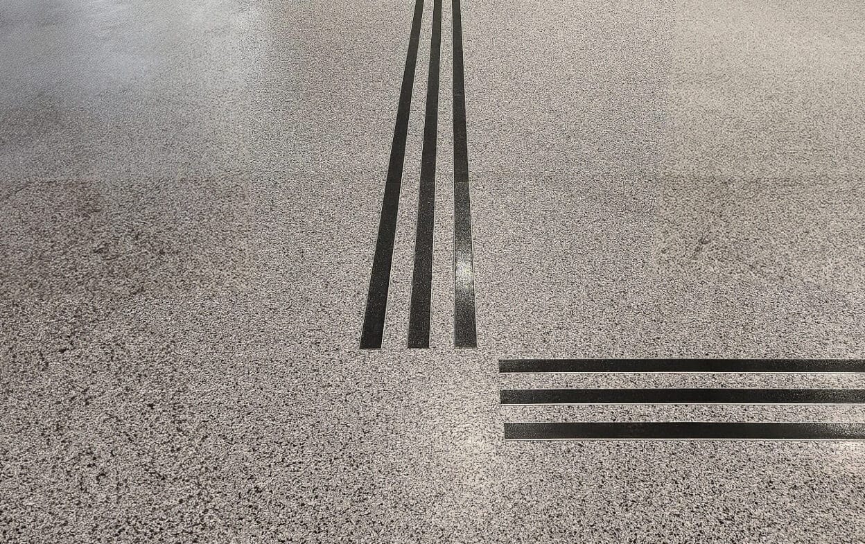 Mörka aluminiumprofiler bildar taktila ledstråk med korsande linjer på ljust terrazzogolv för navigering i offentlig miljö.