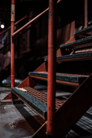industritrappa med svarta halkskyddslister på metalltrappsteg med röda ledstänger i en mörk industrimiljö.