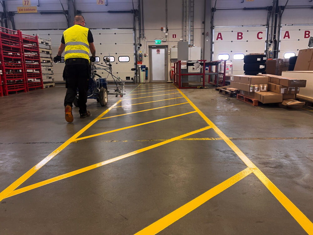 Lagerarbetare i skyddsväst följer gul Safestep Linjemålning med lastningsutrustning i distributionscenter.