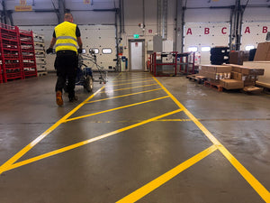 Lagerarbetare i skyddsväst följer gul Safestep Linjemålning med lastningsutrustning i distributionscenter.