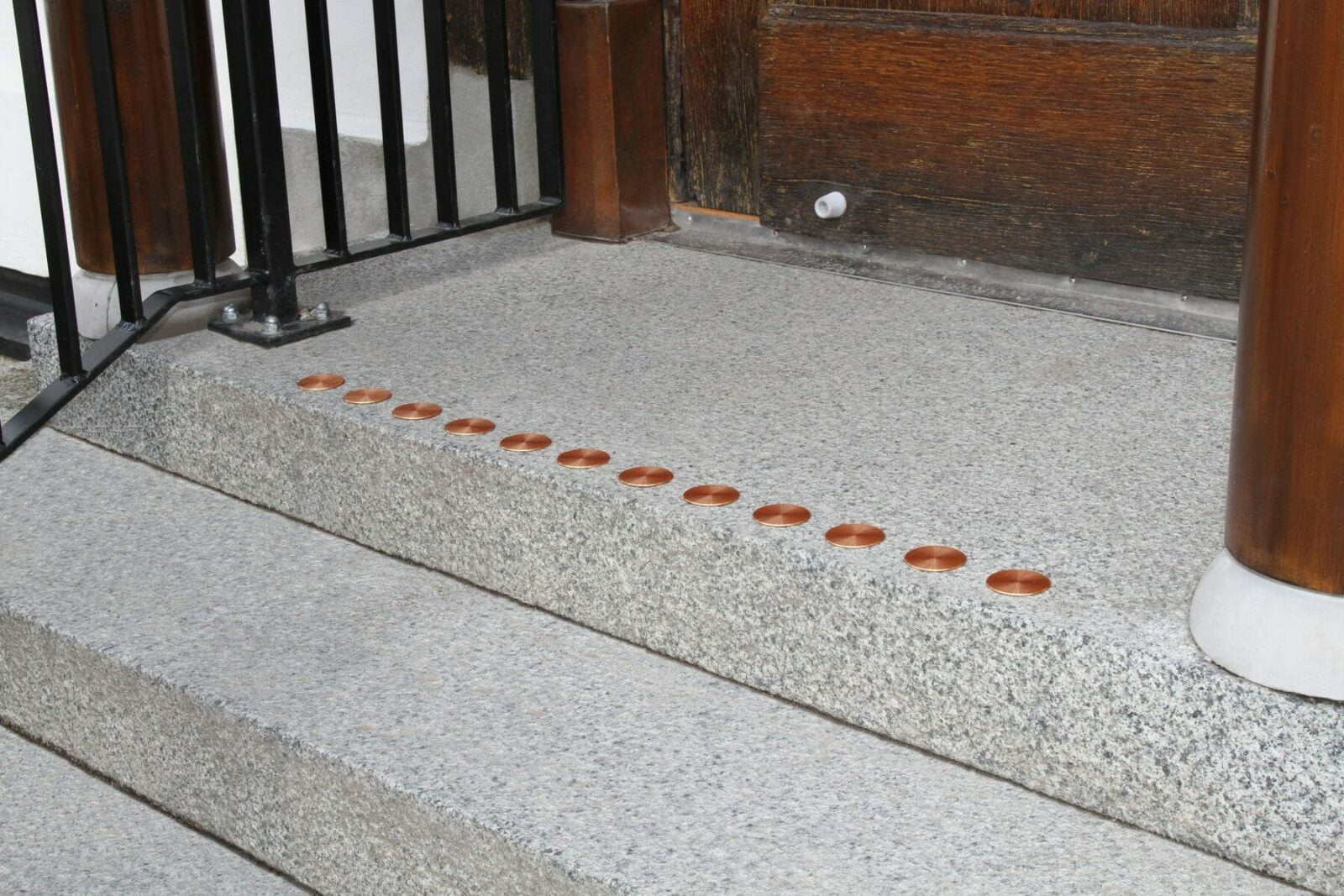 Kontrastmarkeringar i form av kopparfärgade runda indikationer längs kanten av ett betongtrappsteg vid en entré med trädetaljer och svart räcke.