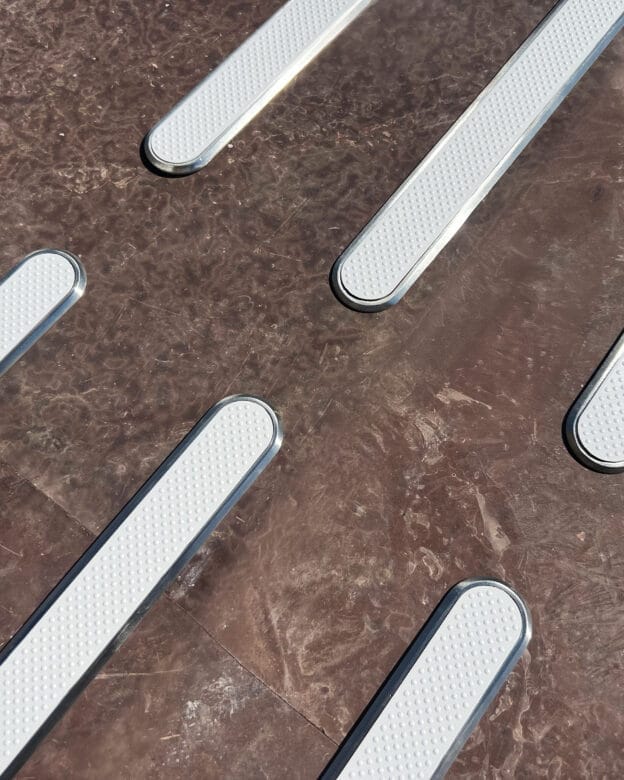 Taktila ledstråk i vitt med upphöjd struktur installerade i en brun golvyta. Ledstavarna är infällda i golvet med metallkanter och har ett texturerat mönster. 