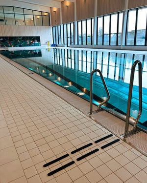 Inomhusbassäng med svarta taktila markeringar på ljust klinkergolv vid poolkanten. Kontrastmarkeringarna varnar för nivåskillnaden och ökar säkerheten i våtmiljön. 