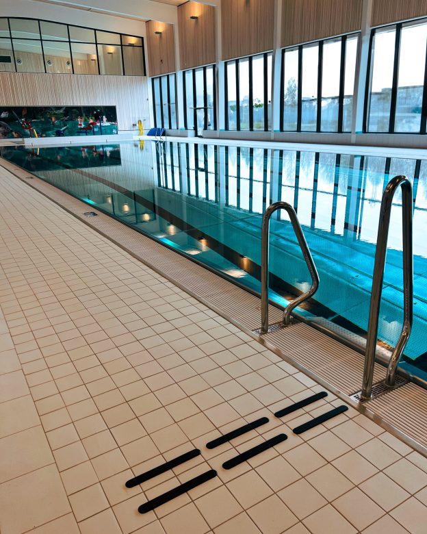 Inomhusbassäng med svarta taktila markeringar på ljust klinkergolv vid poolkanten. Kontrastmarkeringarna varnar för nivåskillnaden och ökar säkerheten i våtmiljön. 