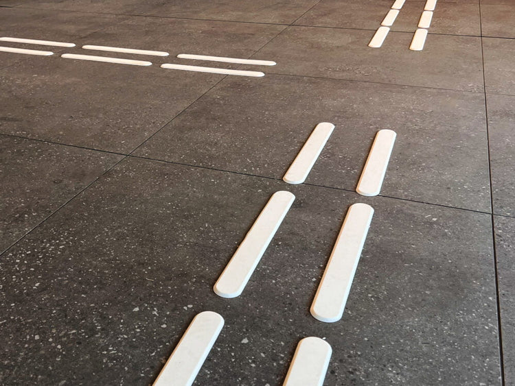 Taktila ledstråk i vit polyuretan monterade på mörkt betonggolv i offentlig miljö. 