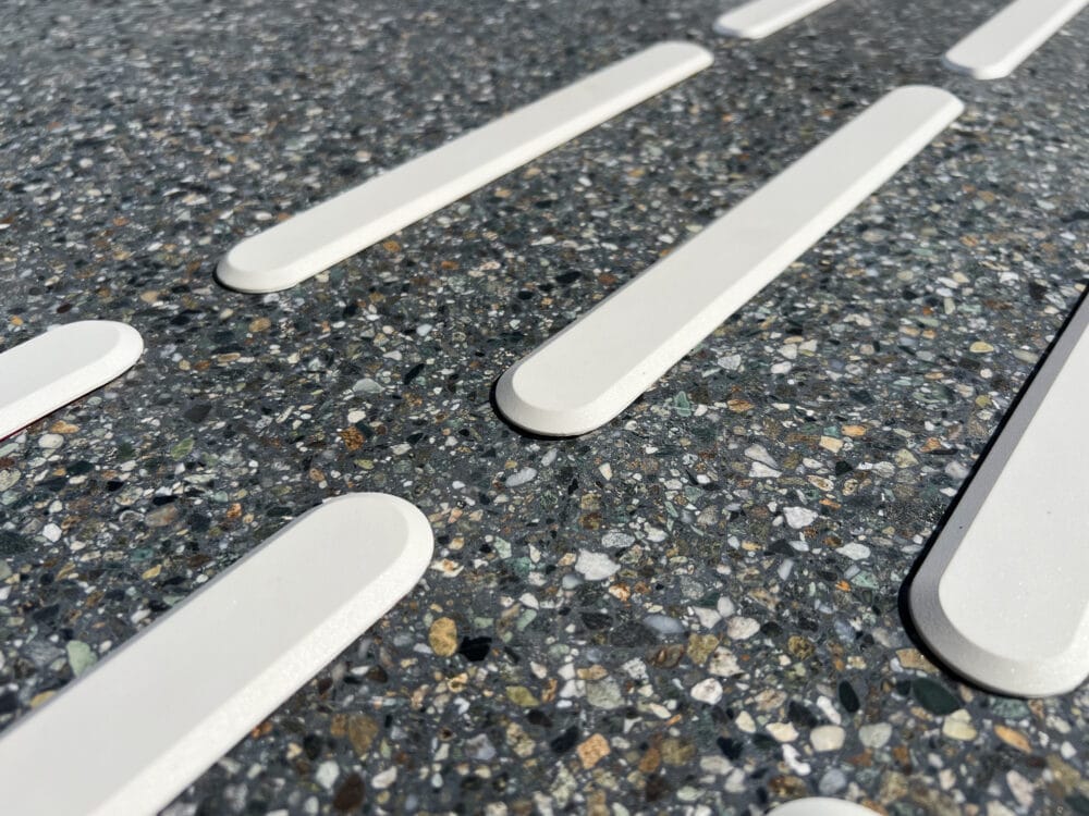 Närbild på vita taktila ledstavar i polyuretan monterade på mörkt terrazzogolv. Markeringarna har en slät, upphöjd yta som ger både visuell kontrast och taktil ledning. 