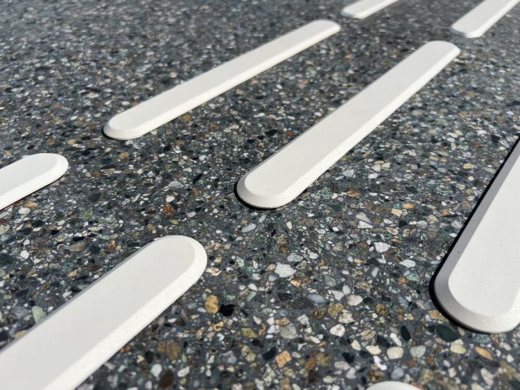 Närbild på vita taktila ledstavar i polyuretan monterade på mörkt terrazzogolv. Markeringarna har en slät, upphöjd yta som ger både visuell kontrast och taktil ledning. 