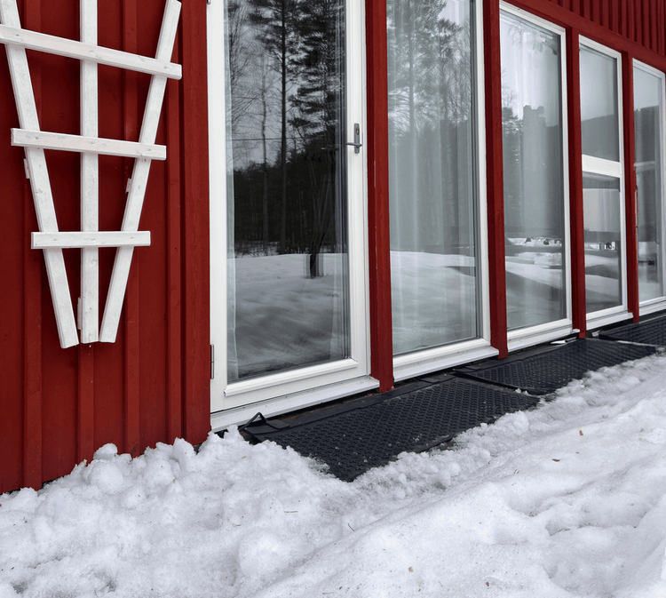 Svarta värmemattor placerade som dörrmatta utanför glasdörrar på en röd stuga.