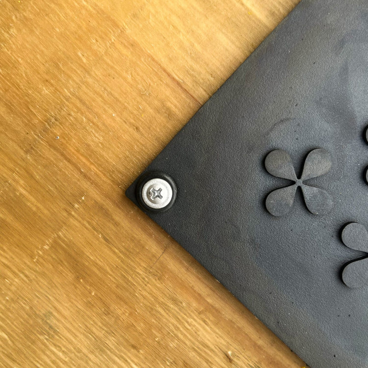 Svart gummipackning med blomformade utskärningar och metallskruv för montering av Safestep värmematta på träunderlag.